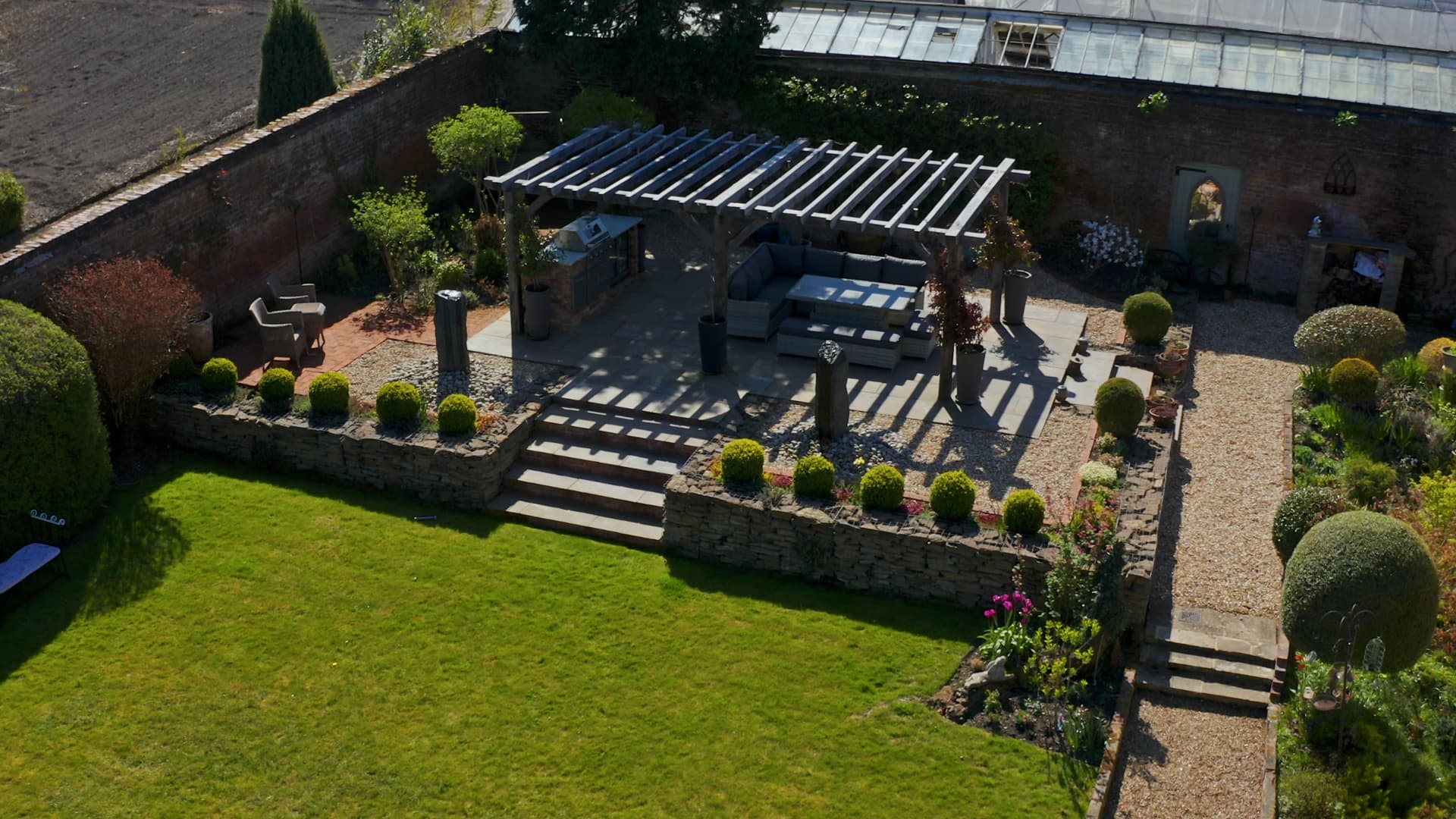 An elevated garden terrace with a pergola, outdoor kitchen, seating and manicured lawns